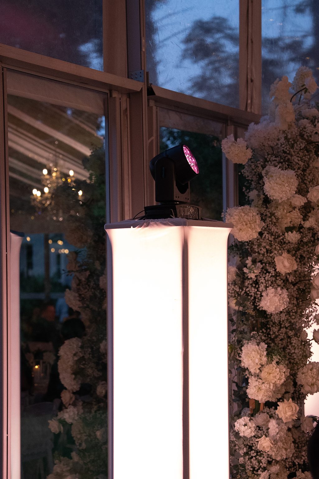 A moving-head lighting fixture glowing on a lit pedestal beside a floral arch
